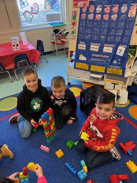 Three young boys are playing with colorful building blocks on a blue carpet in a classroom.