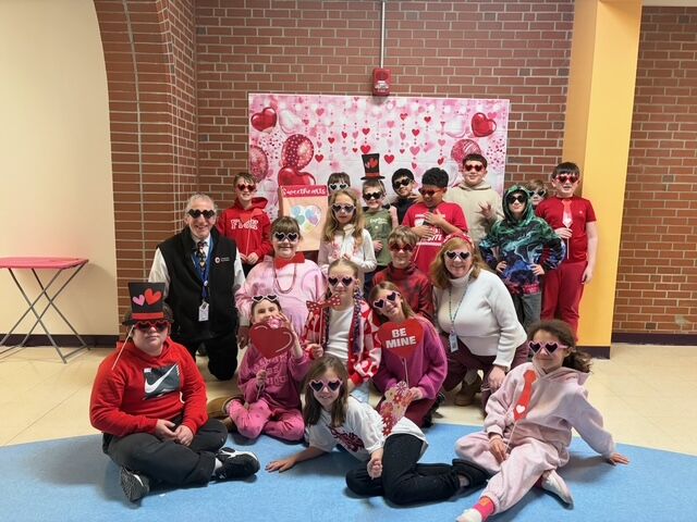 A group of students and adults pose for a photo in front of a Valentine's Day themed backdrop, all wearing heart-shaped glasses.