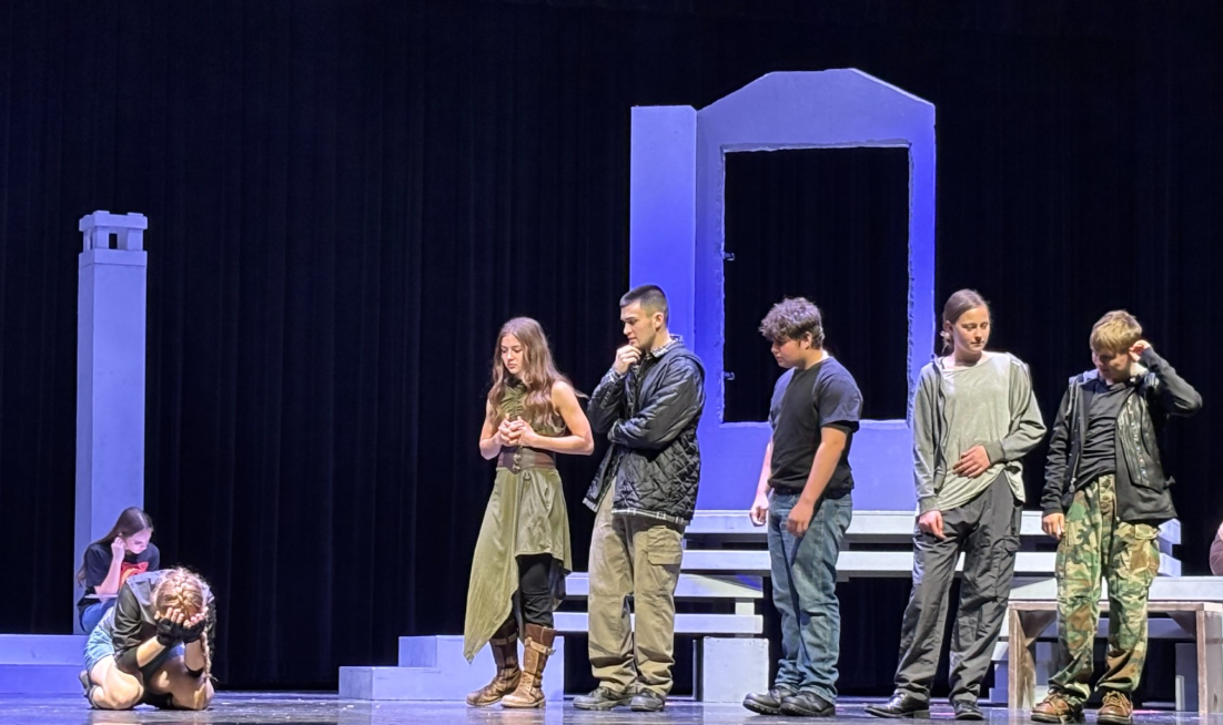 A group of young actors stand on a stage with theatrical props, including a large frame and a pillar.