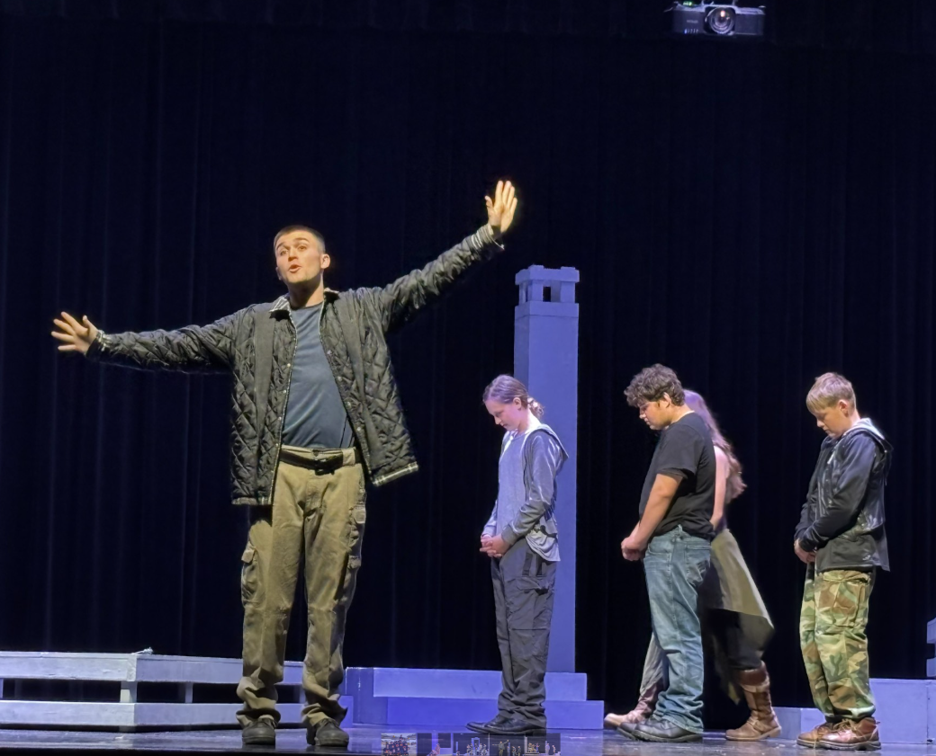 A young man on stage gestures with open arms, addressing an audience while other performers stand with heads bowed.