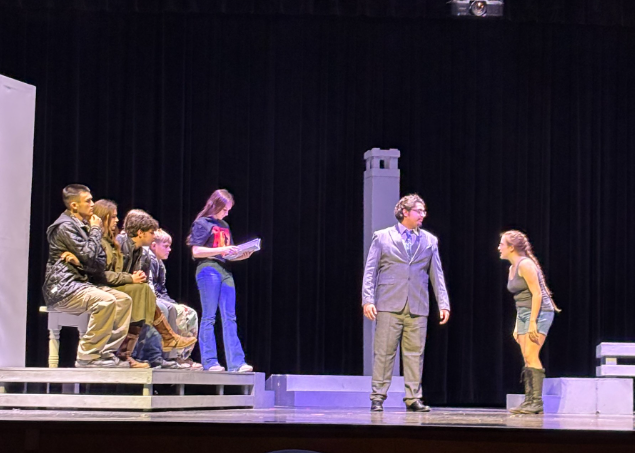 A group of young actors rehearse a scene on a stage with a black curtain backdrop.
