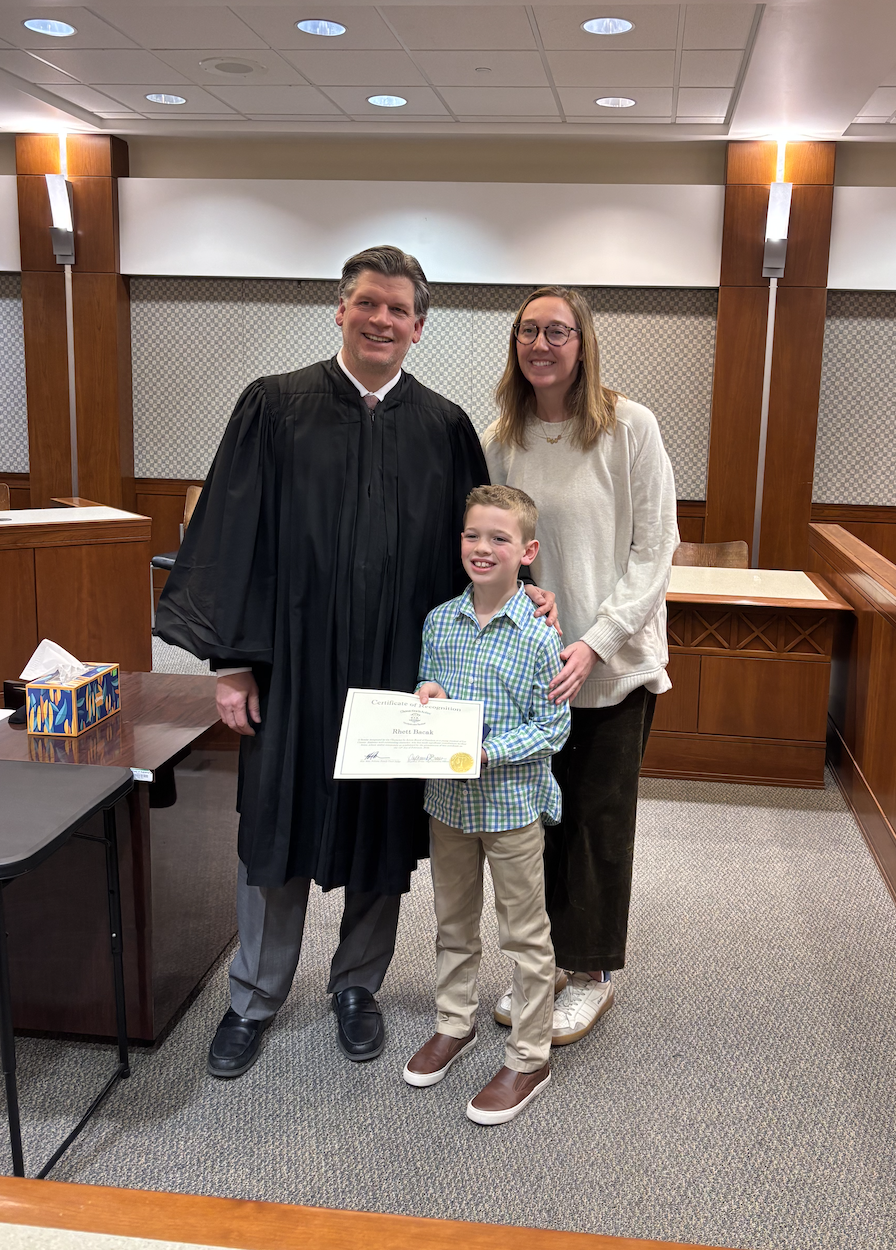 A judge in a black robe stands with a smiling boy holding a certificate and a woman in a courtroom.
