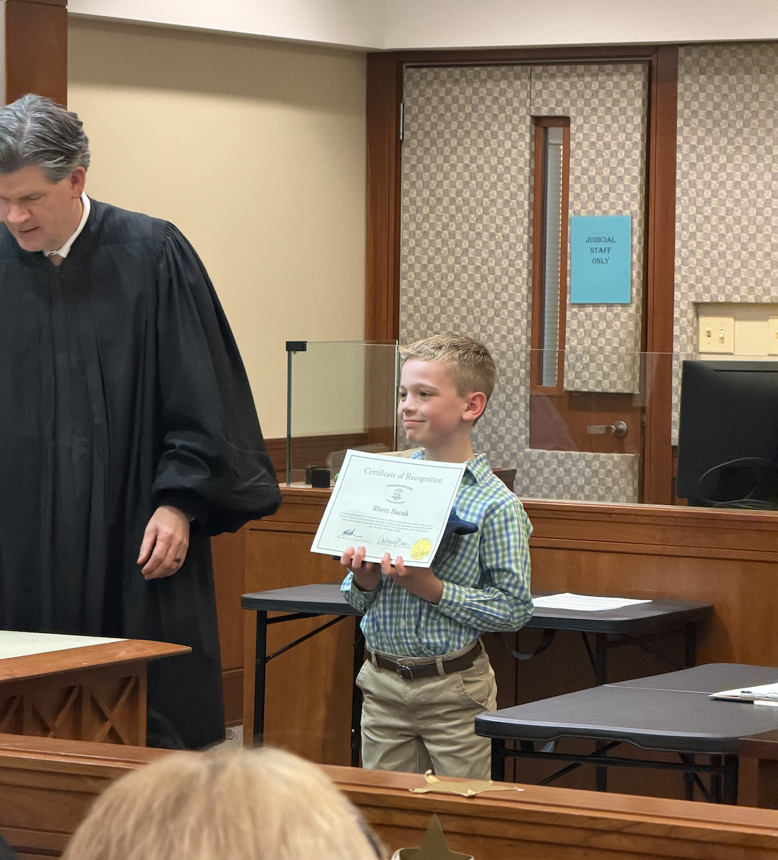 A young boy in a plaid shirt proudly holds a "Certificate of Recognition" in a courtroom setting.