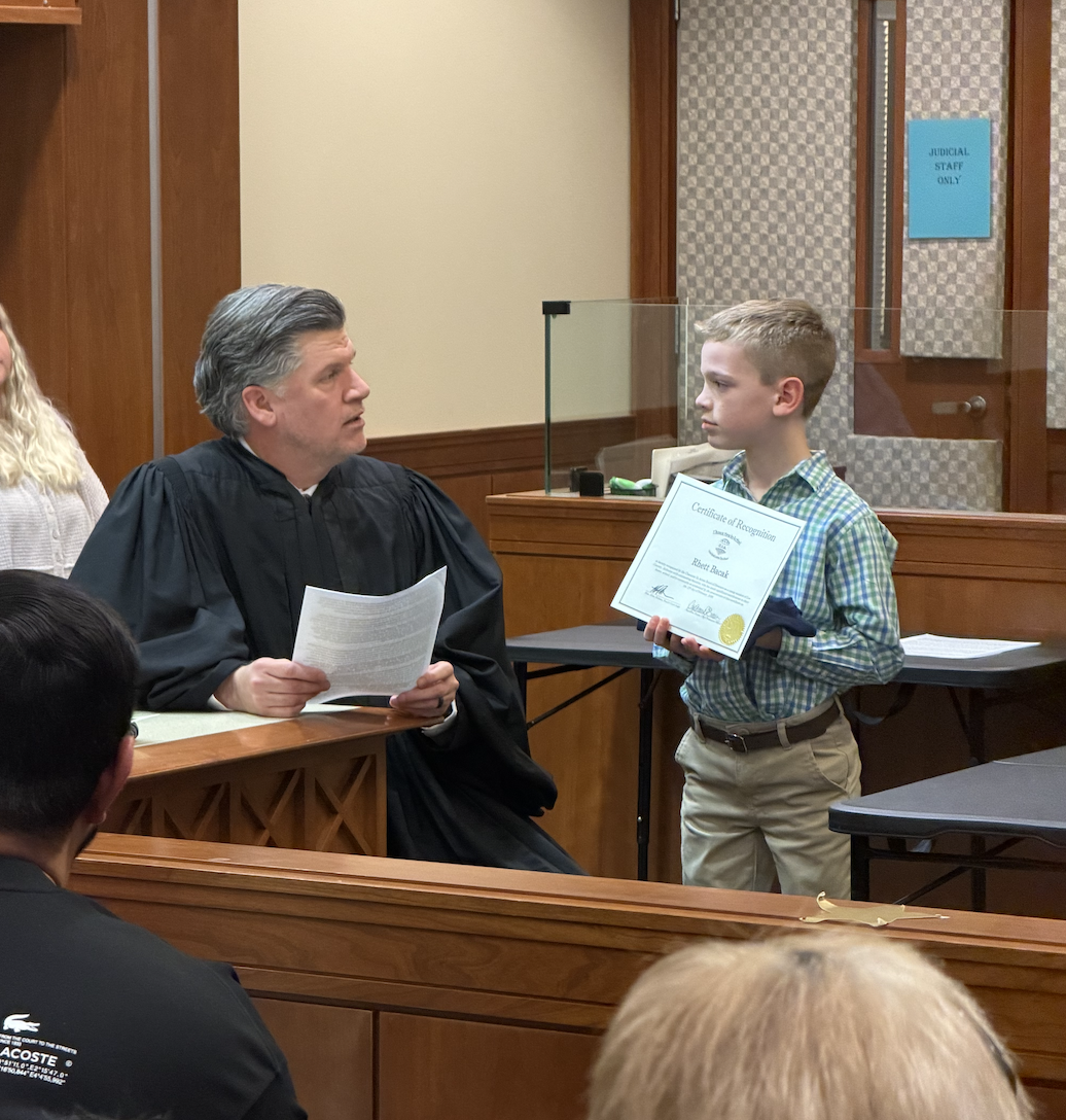 A judge in a black robe holds papers while speaking to a young boy holding a Certificate of Recognition.