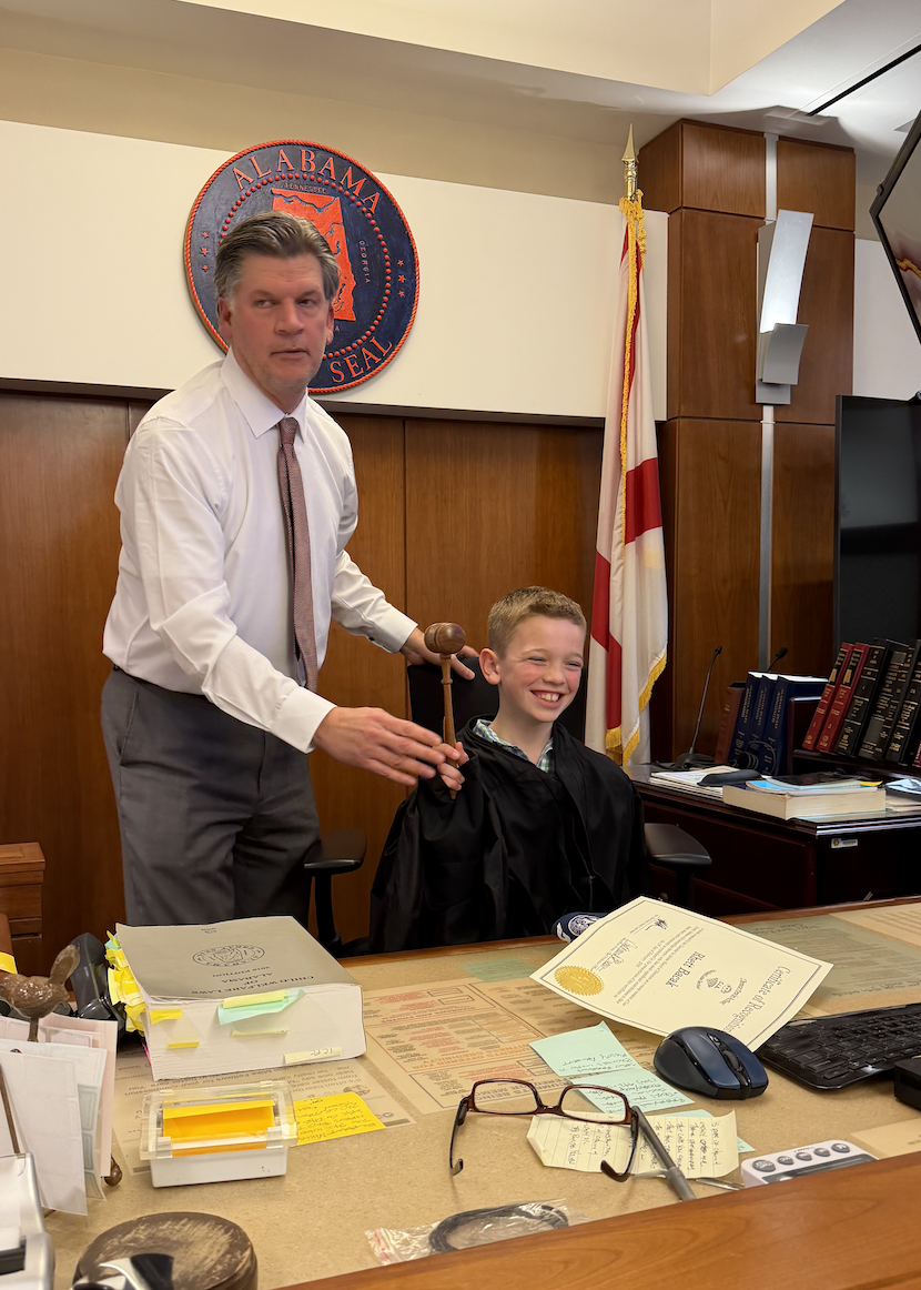 A smiling boy in a judge's robe sits in a courtroom, with an adult holding a gavel behind him.