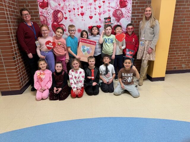Two teachers and a group of children pose for a Valentine's Day photo in a classroom decorated with hearts.