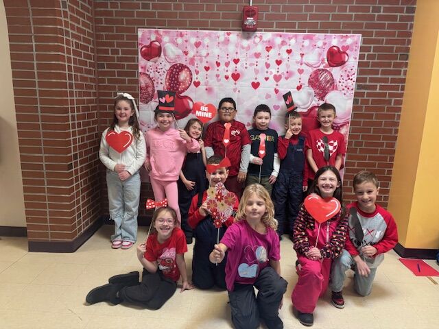 A group of children pose for a Valentine's Day photo in front of a heart-themed backdrop.