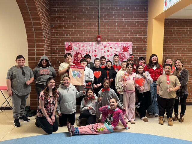 A group of students and a teacher pose for a photo in front of a Valentine's Day themed backdrop.