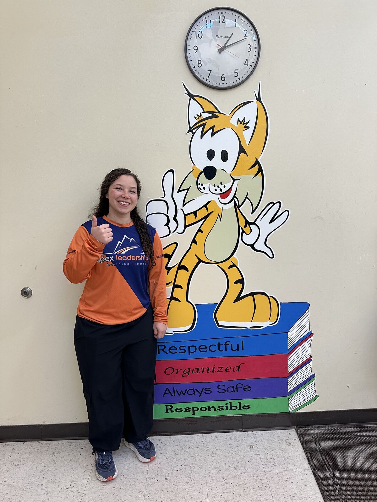 A smiling young person stands next to a cartoon tiger mural and a stack of colorful books with positive character traits.