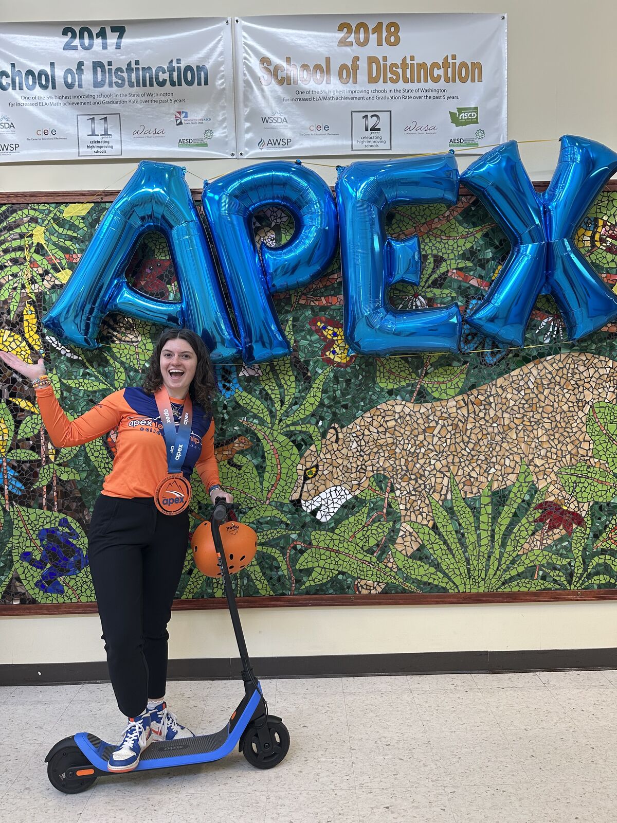 A smiling person in an orange shirt and black pants stands on a blue electric scooter, with large blue balloons spelling 'APEX' behind them.