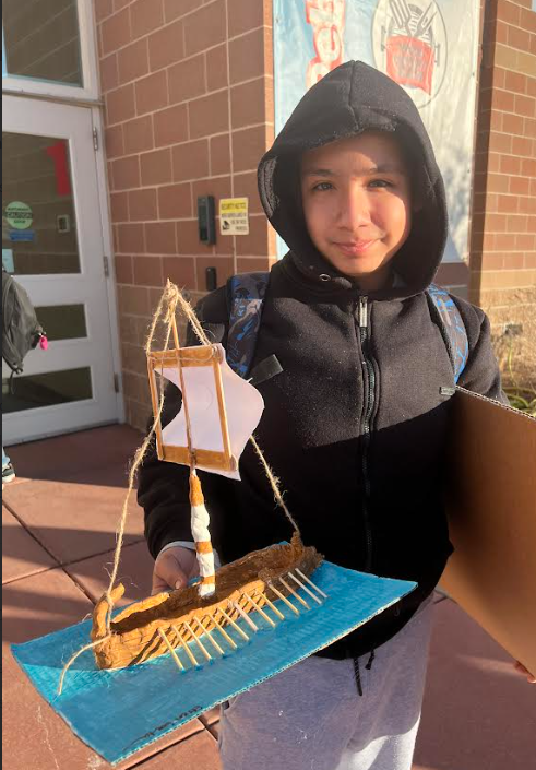 A young person proudly displays a handmade model of an ancient ship with sails.