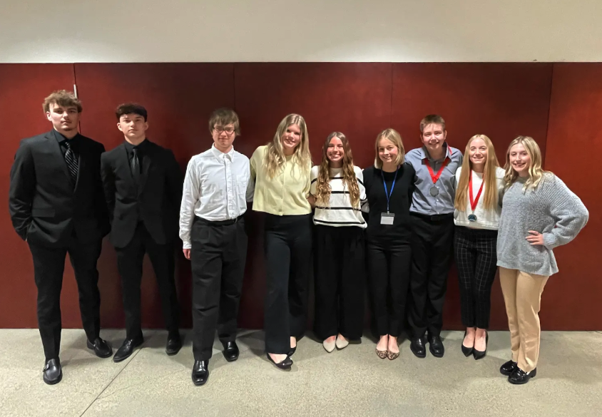 A group of nine young people stand together in front of a maroon wall.