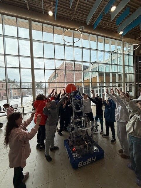 A group of students surrounds a tall, multi-level robot with an orange ball on top, raising their hands.