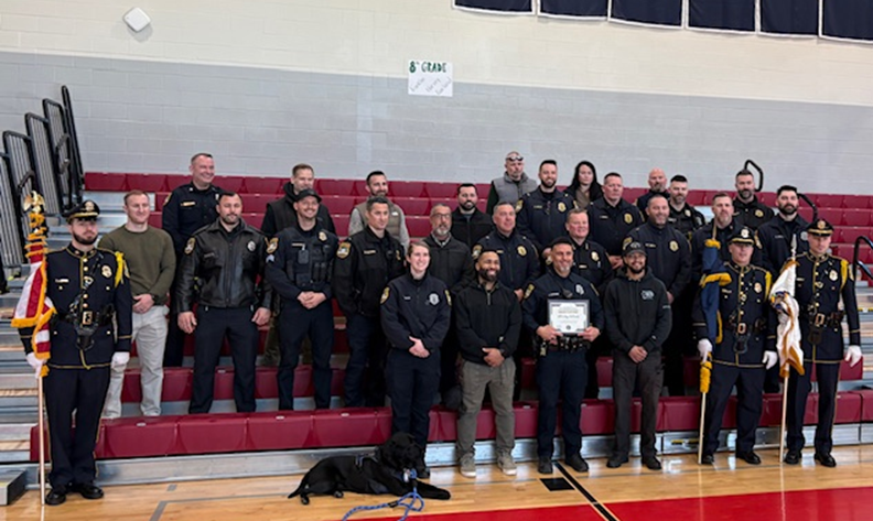 A group of police officers and civilians pose for a photo in a gymnasium with bleachers.