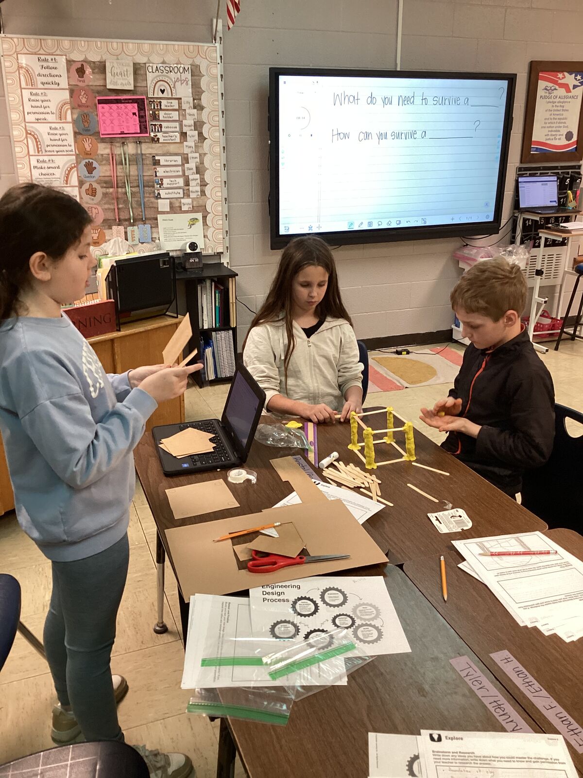 Students work on a STEM project, building with craft sticks and clay, with a digital display showing "What do you need to survive a ?" and "How can you survive a ?".