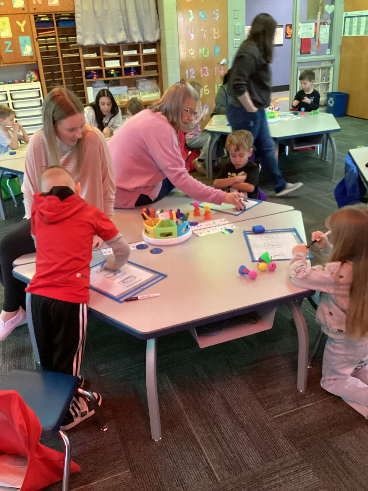 A teacher guides young students in a classroom activity at a table with colorful manipulatives.