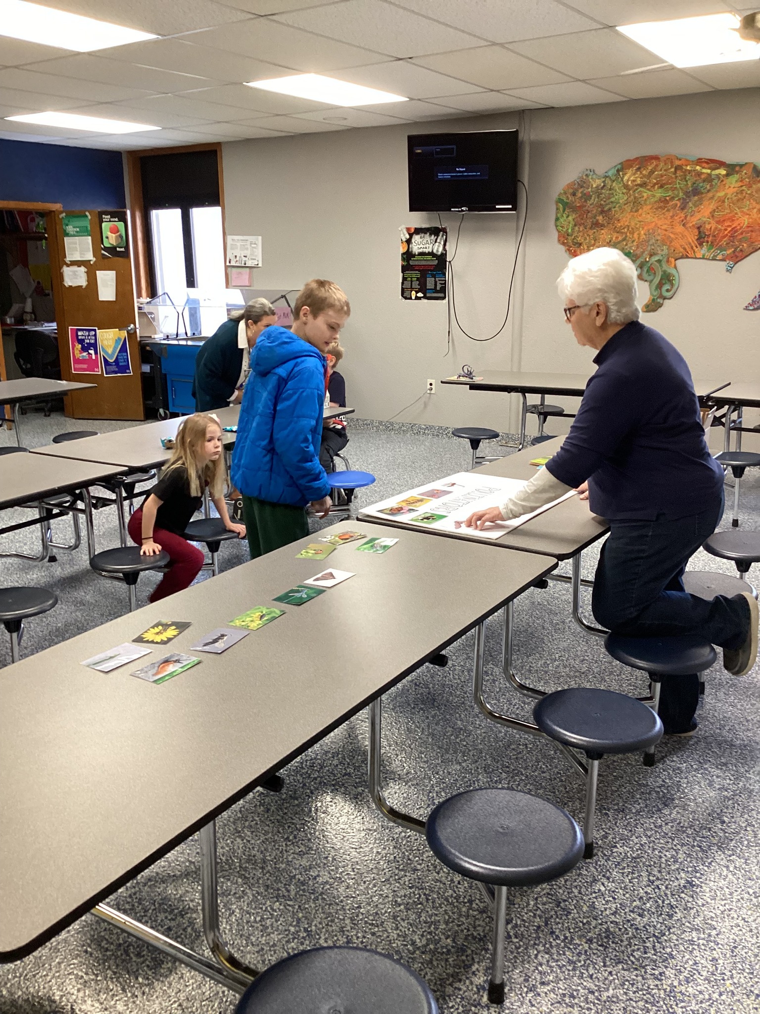 An adult instructor guides children in a classroom setting, pointing to a poster on a table.