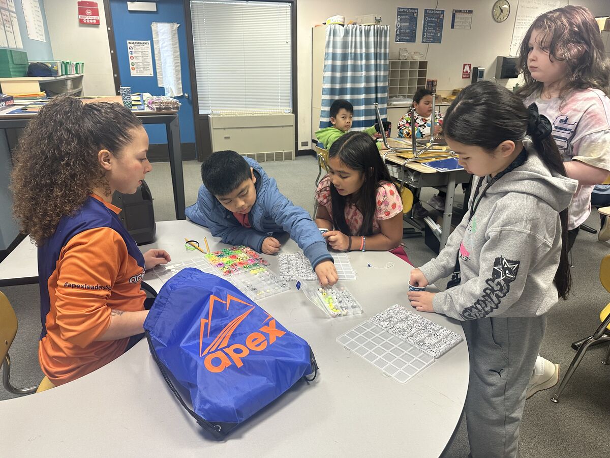 Children and an adult are gathered around a table in a classroom, working with beads and string.