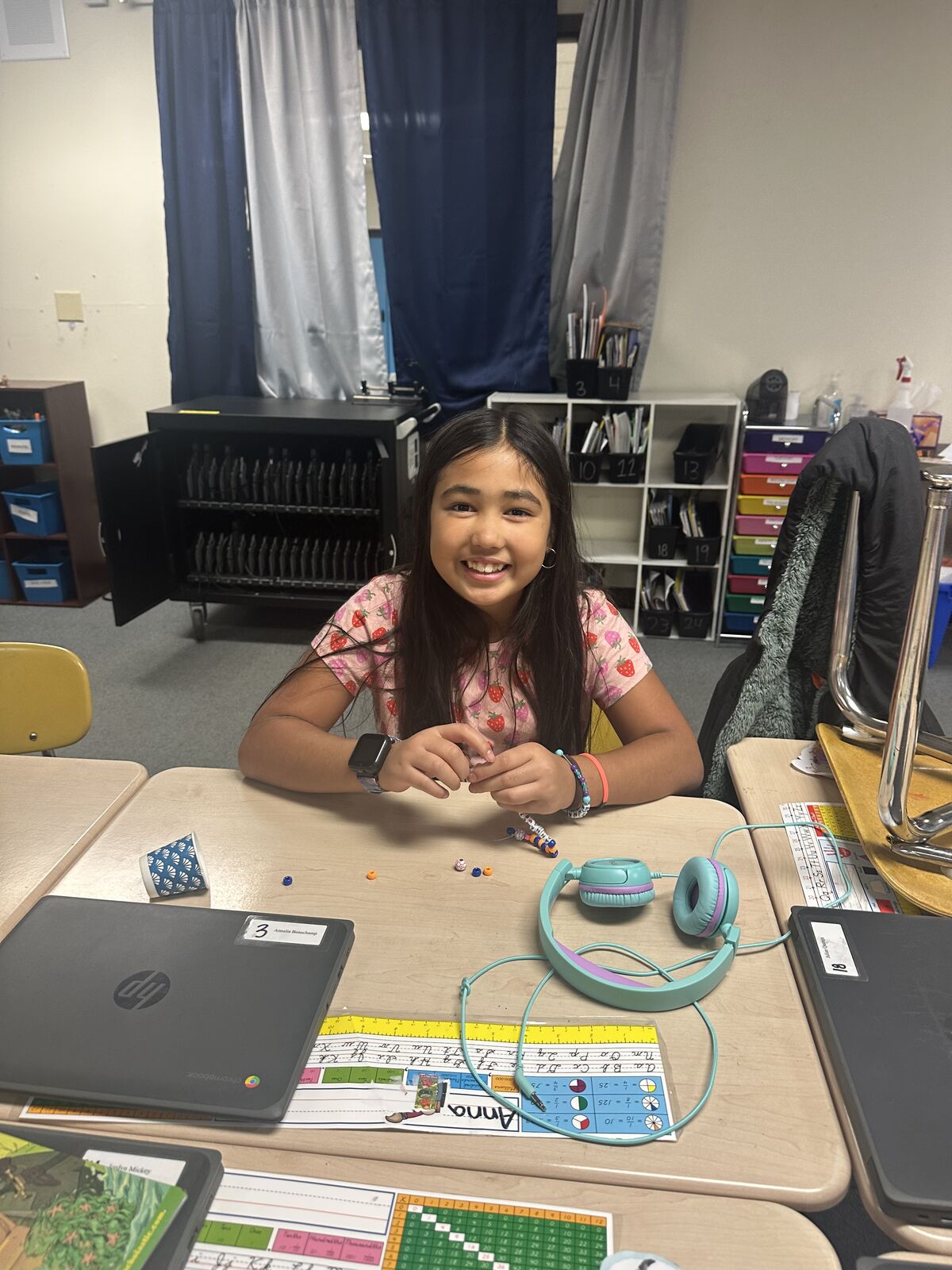A young girl with long dark hair smiles while sitting at a desk, working with colorful beads.