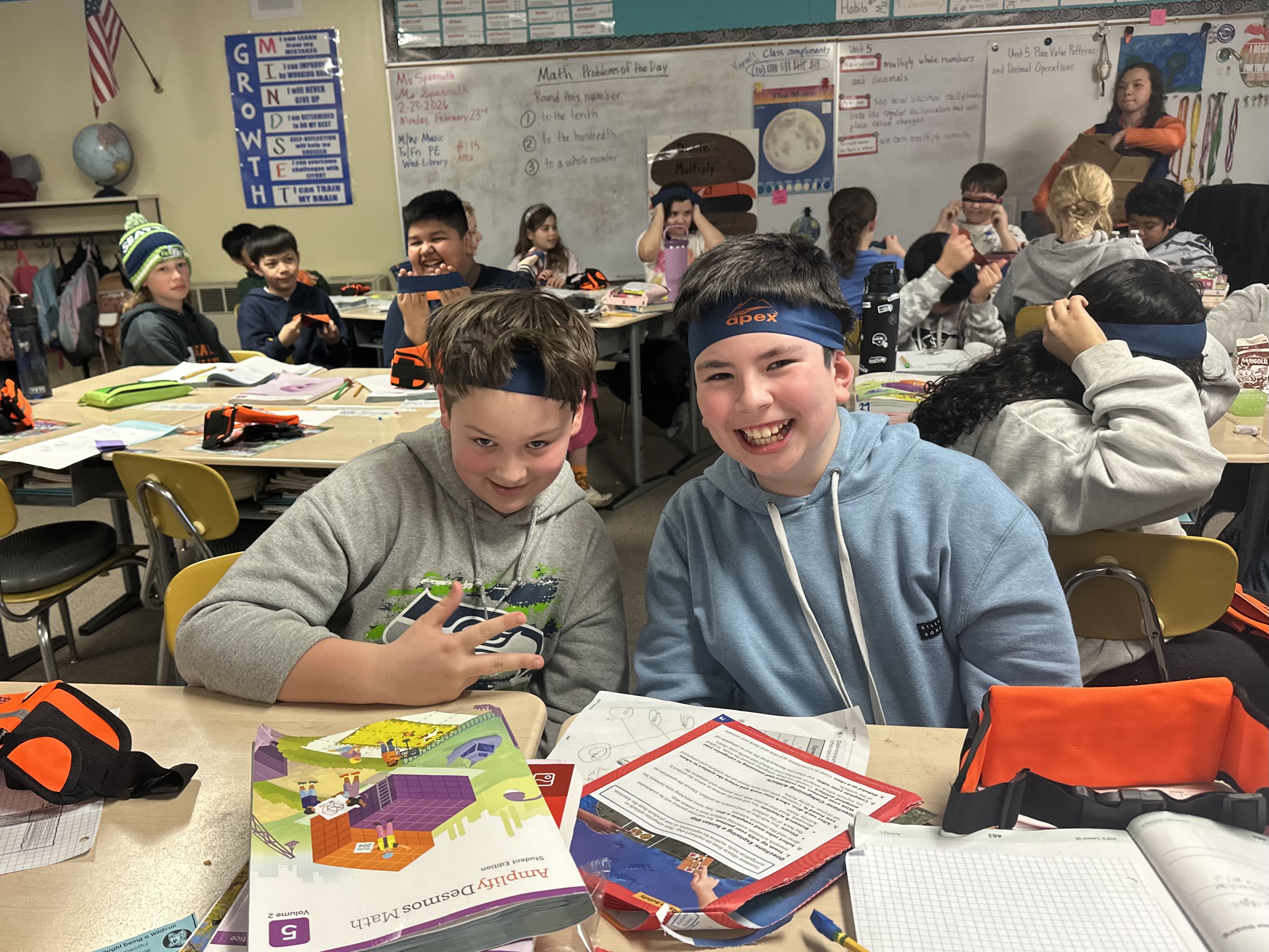 Two smiling boys in a classroom wear headbands, one with the word 'apex' visible, while other students are in the background.