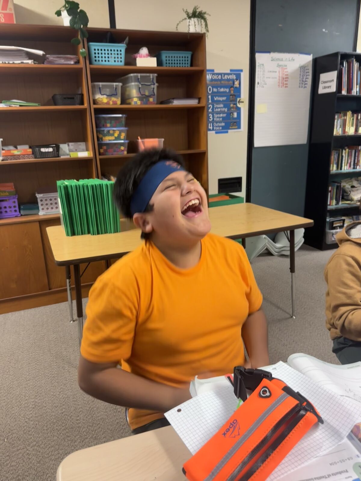A young student in an orange shirt and blue headband laughs heartily in a classroom setting.