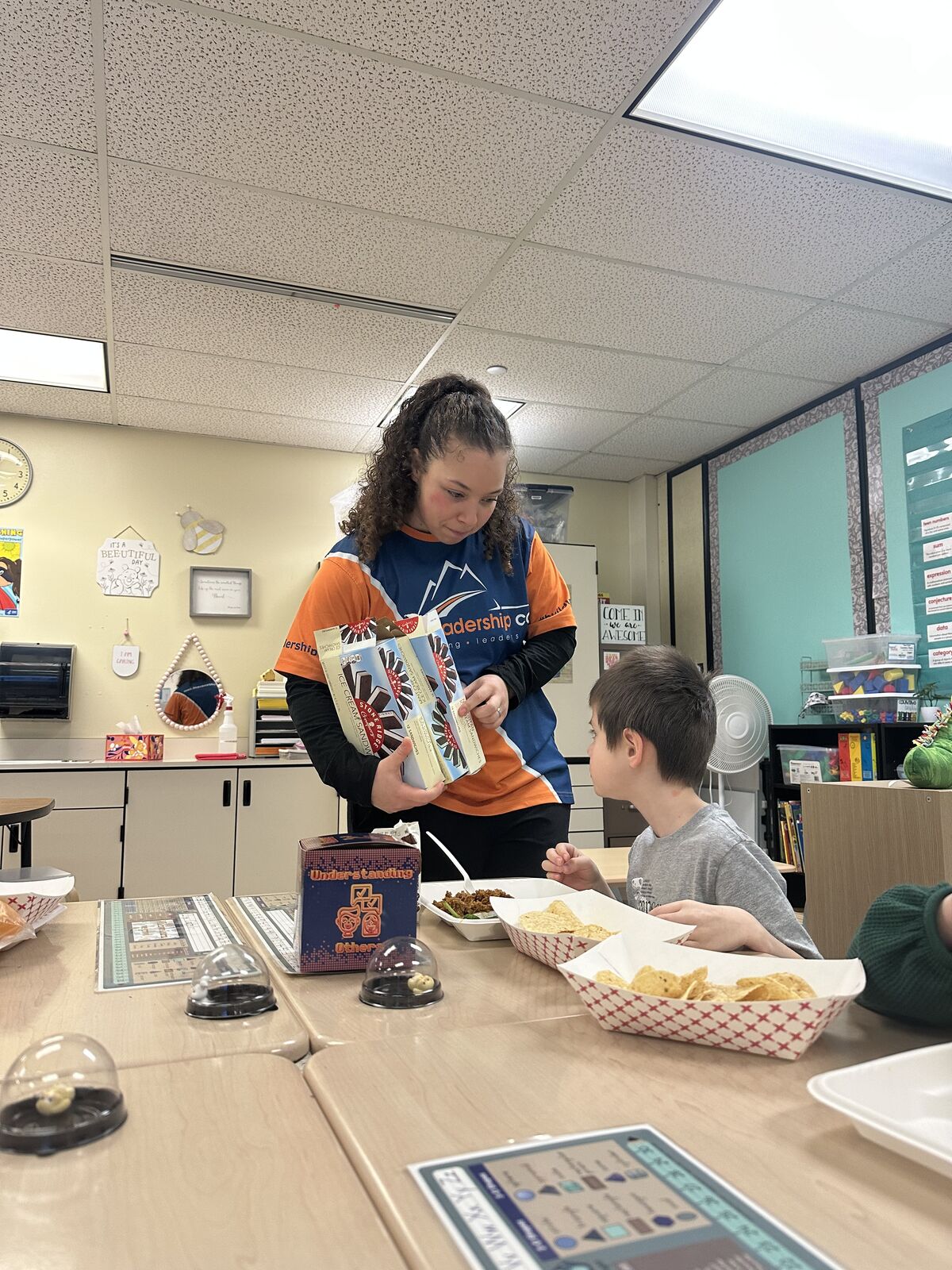 A young girl in an orange and blue shirt holds boxes of ice cream sandwiches while a boy reaches for chips.