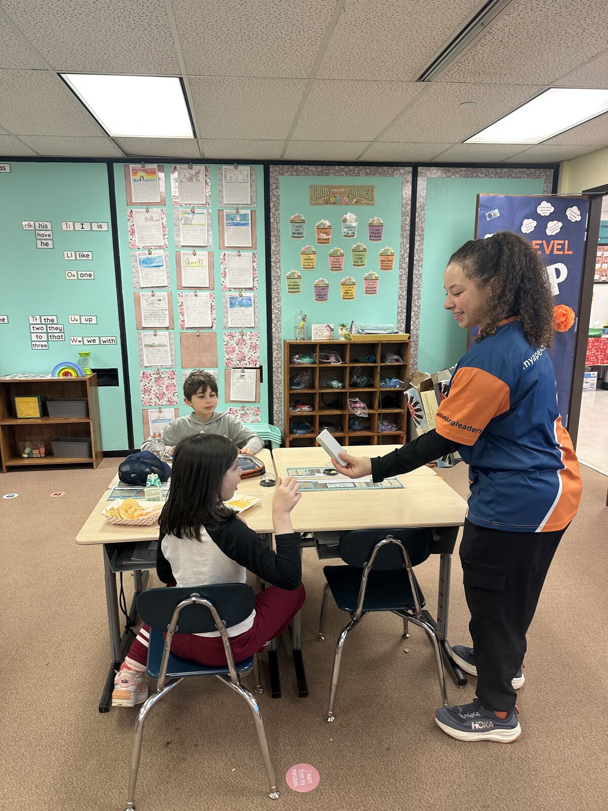 A teacher in an orange and blue shirt interacts with students at desks in a classroom.