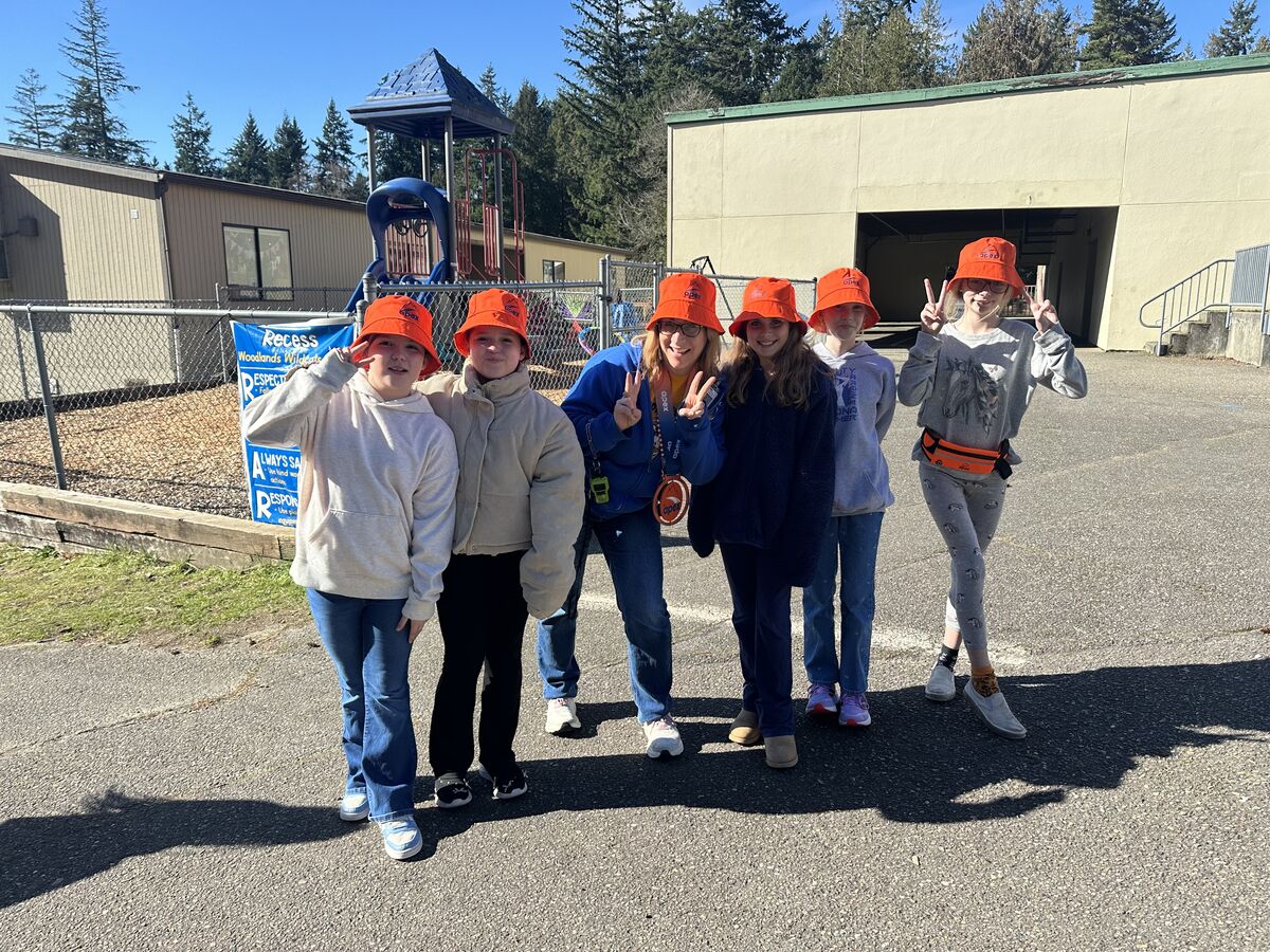 A group of children and an adult wearing orange bucket hats stand together outdoors, smiling and making peace signs.