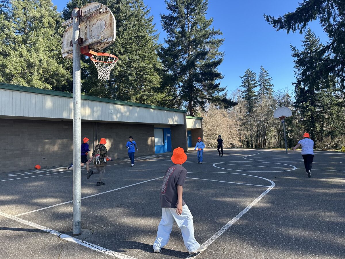 Children in orange hats play basketball on an outdoor court with trees in the background.