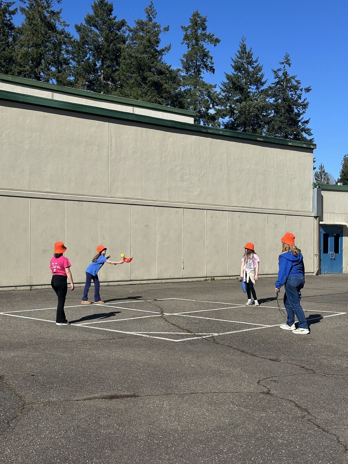 Children in orange hats play on a paved schoolyard with a building and trees in the background.