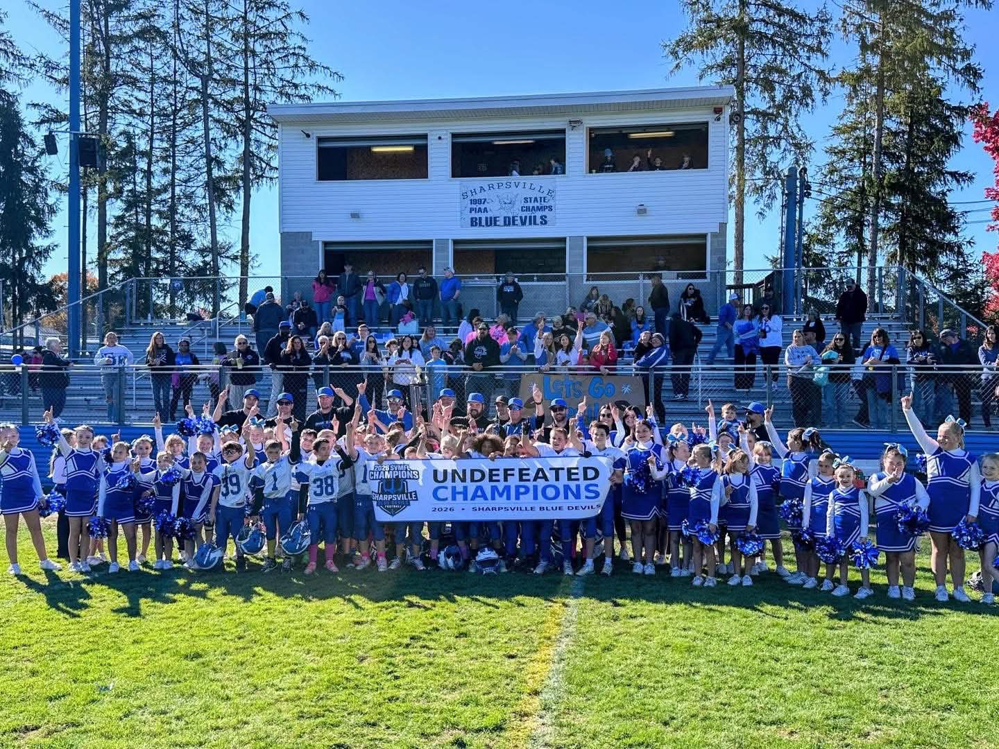 A large group of young football players and cheerleaders in blue and white uniforms celebrate on a grassy field, holding a banner that reads "Undefeated Champions."
