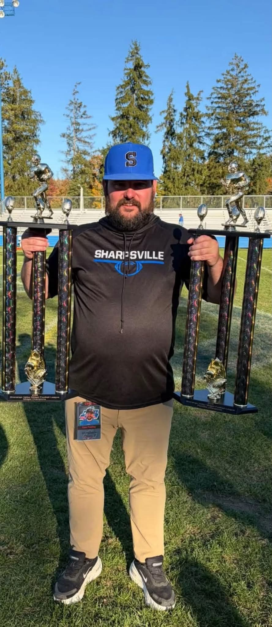 A smiling man wearing a Sharpville hoodie and blue baseball cap proudly holds two large football trophies outside on a sunny day.