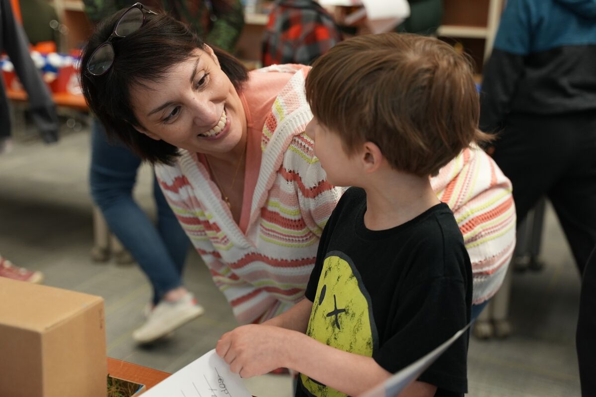 A woman smiles warmly at a young boy holding papers, possibly in a classroom or event.