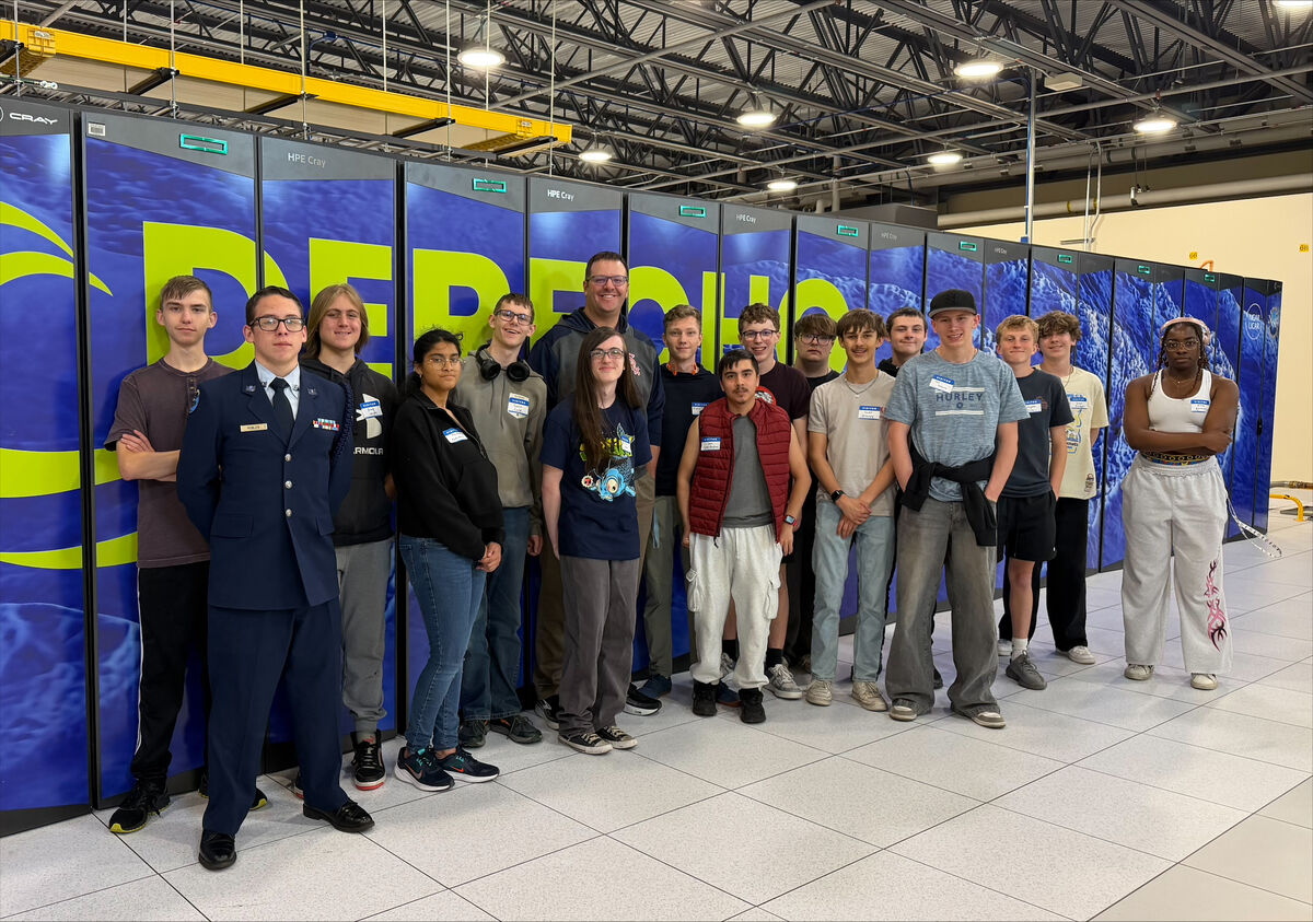 A diverse group of students and adults pose for a photo in front of large blue supercomputer cabinets.