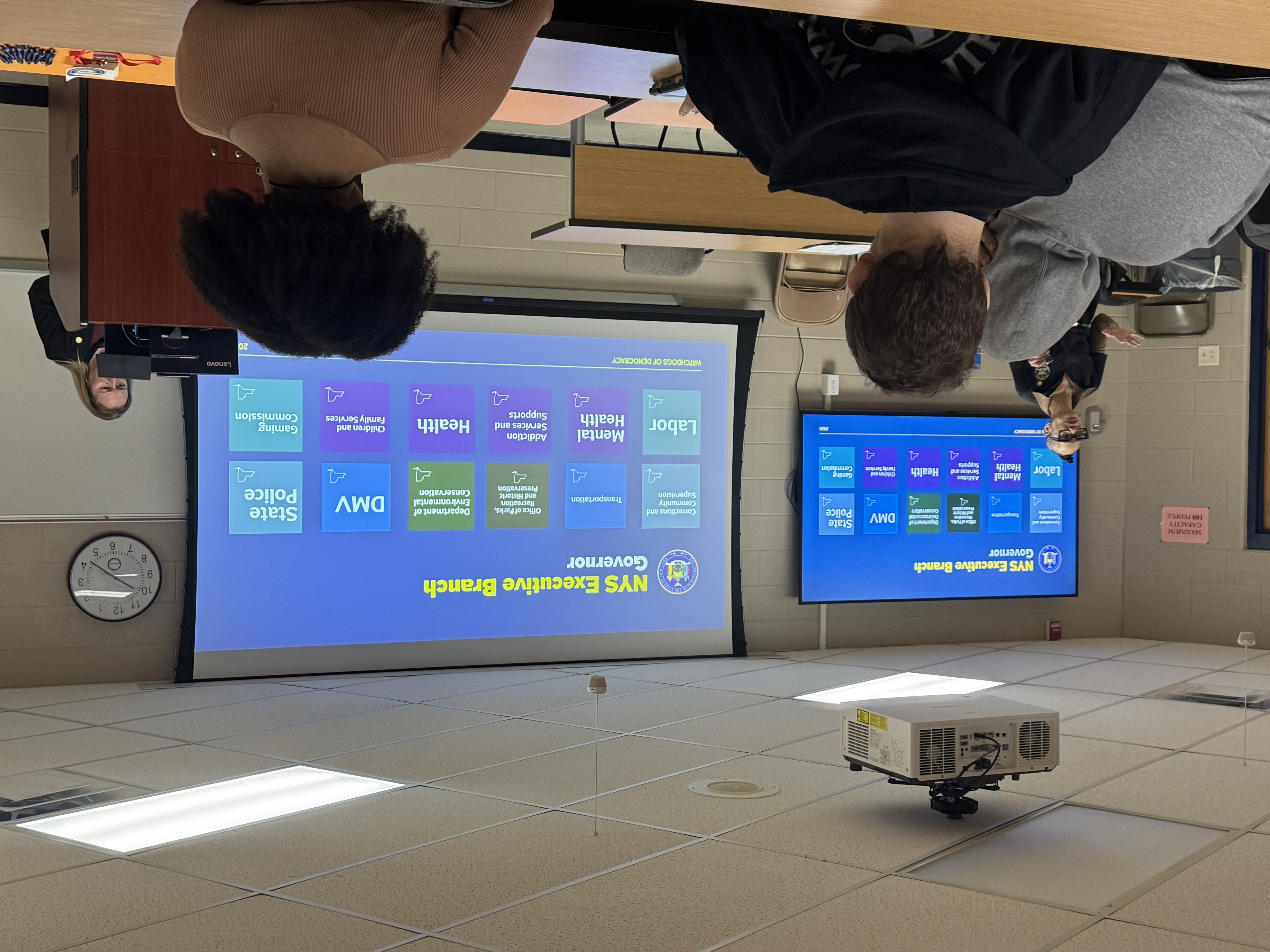 A classroom setting with two large screens displaying information about the NYS Executive Branch and Governor.