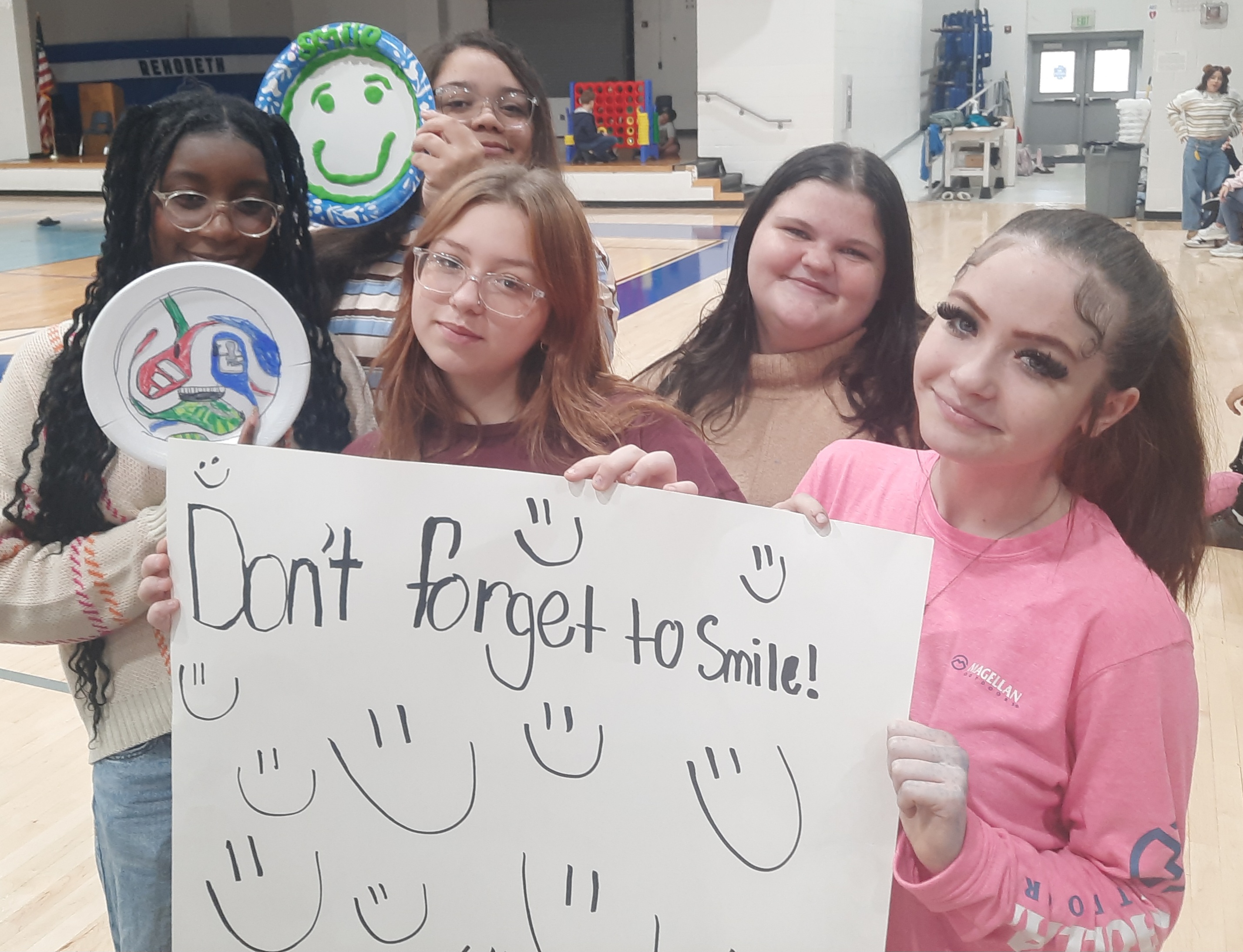 Five smiling young women pose together in a gymnasium, holding a sign that reads "Don't forget to Smile!".