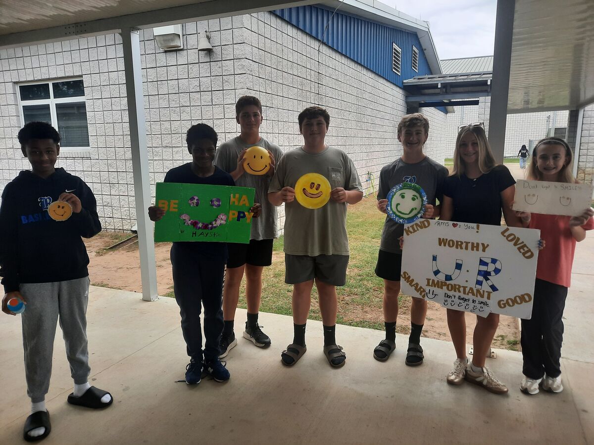 A group of students stand together outside a school building, holding up positive signs and smiley face decorations.