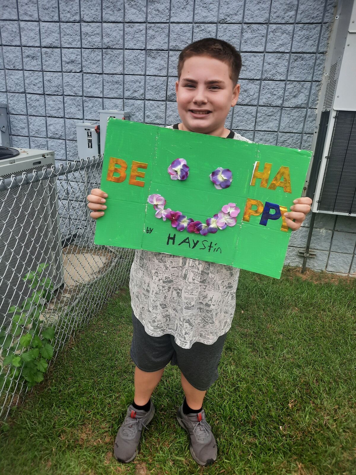 A smiling child holds a handmade green sign that reads "BE HAPPY" with a flower smile.