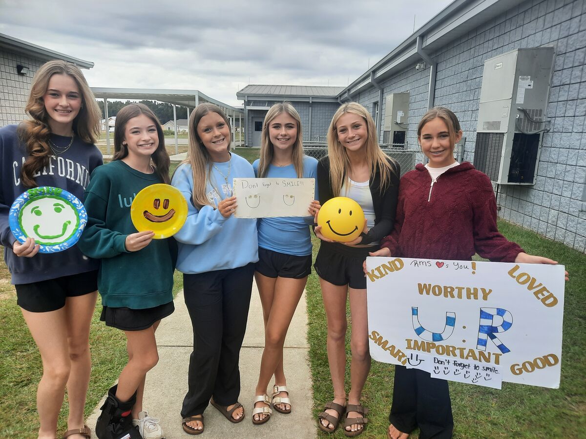 Six smiling young people stand outside a school building, holding smiley face decorations and positive signs.