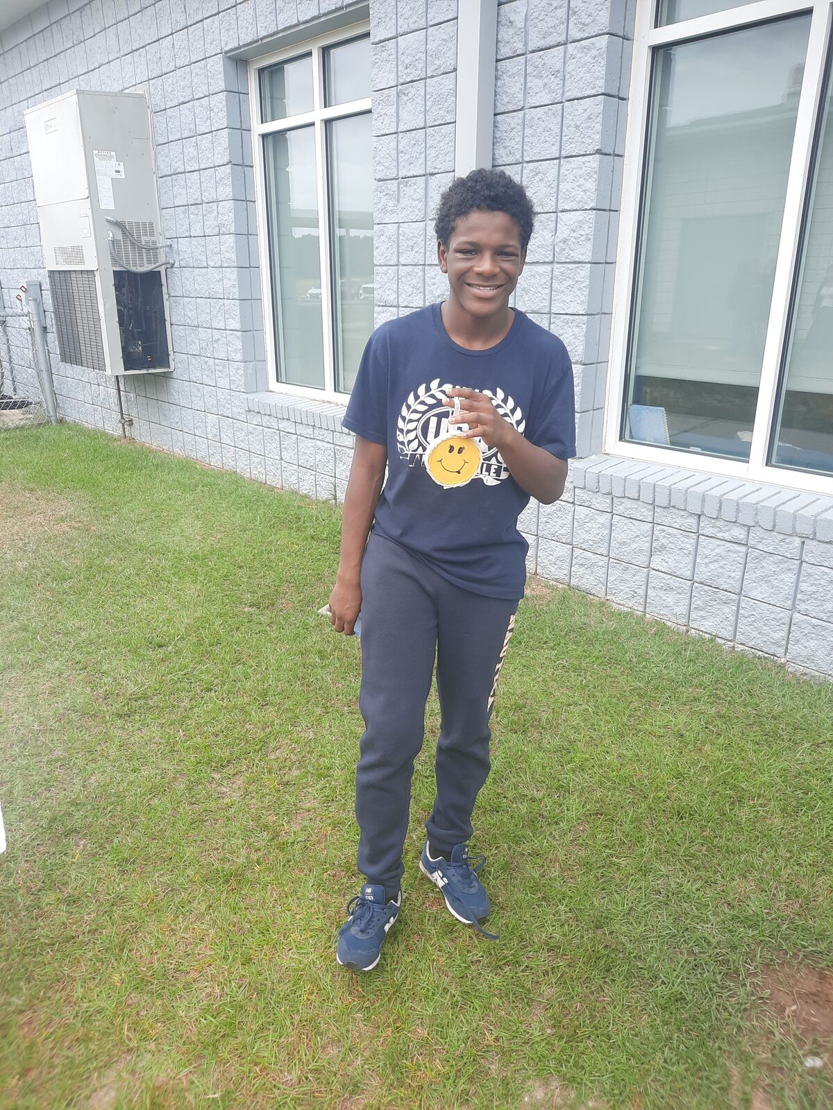 A smiling young person stands on the grass next to a grey brick building.
