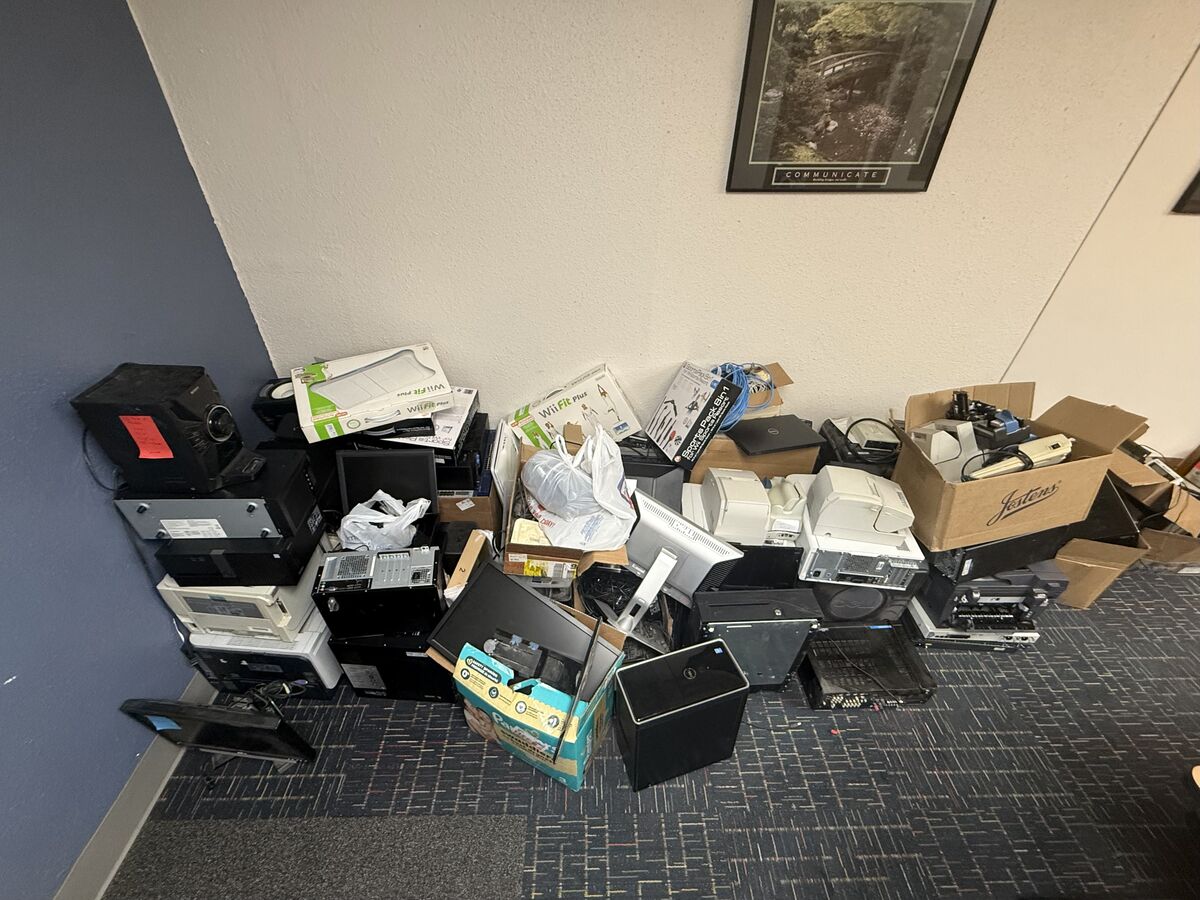 A large pile of discarded electronics, including computer towers, monitors, and printers, is stacked on a carpeted floor.