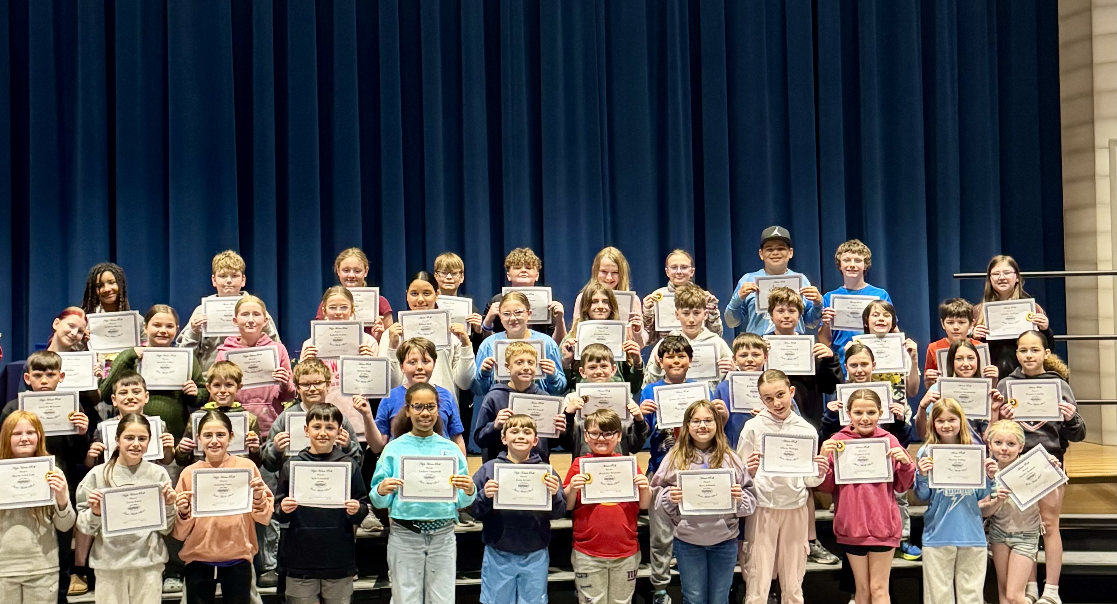 A diverse group of students stands on risers, proudly holding certificates.