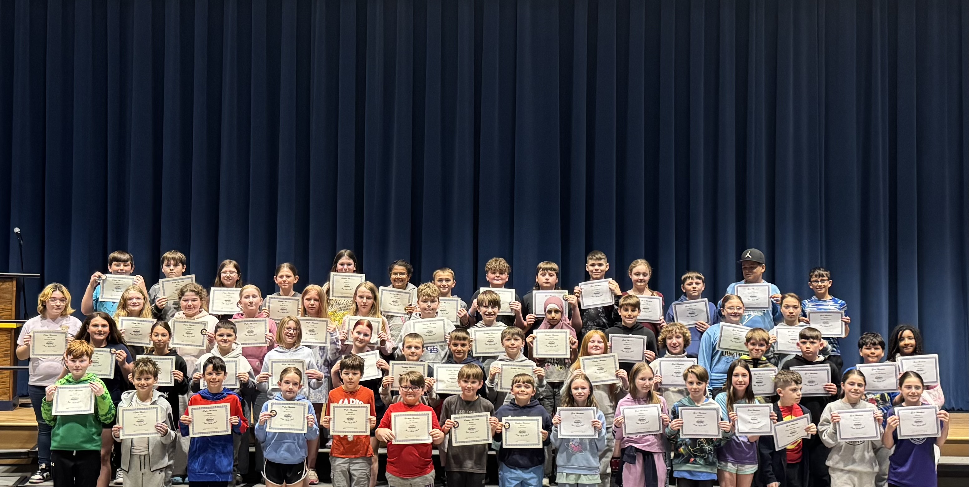 A large group of students stands on risers, holding certificates in front of a blue curtain.