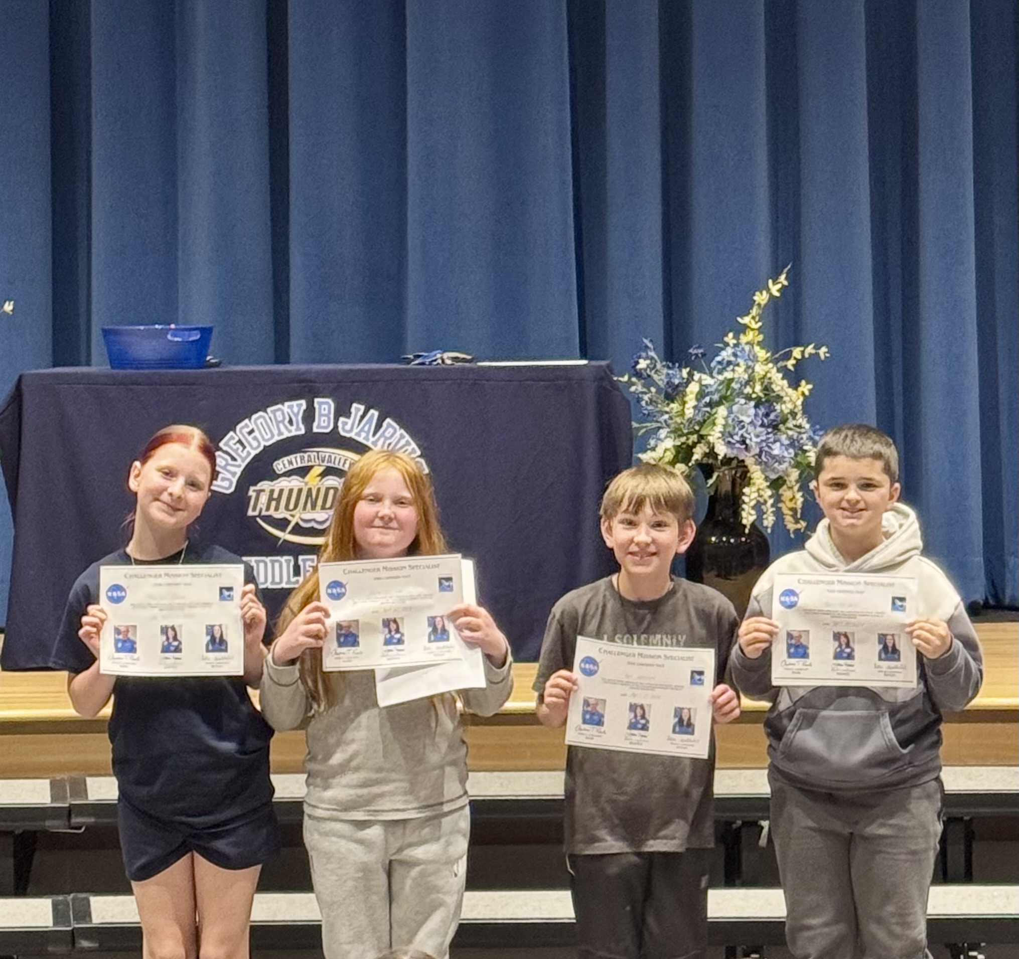 Four students proudly hold certificates from a NASA-related program, standing on a stage with a blue backdrop.