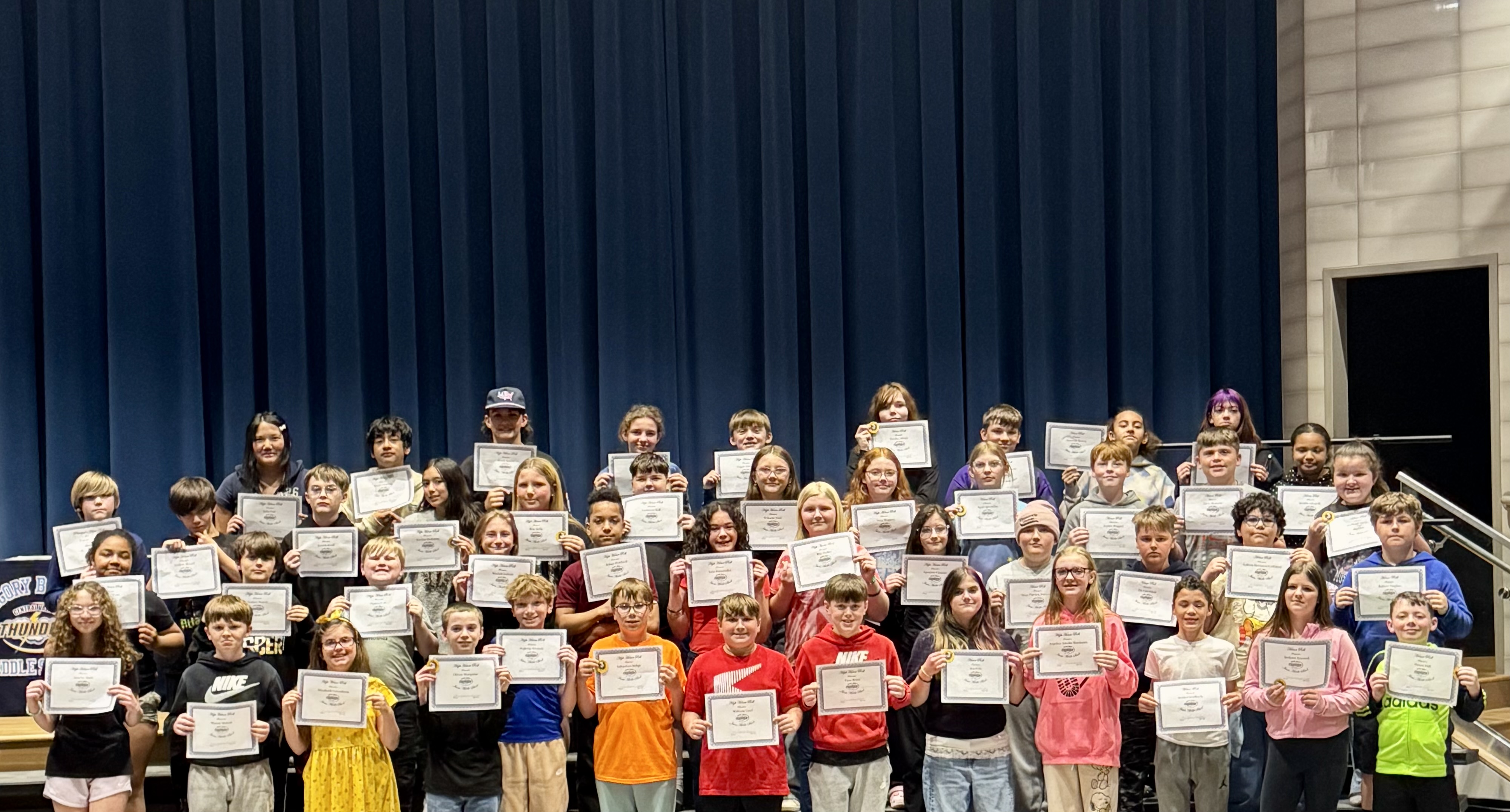 A diverse group of students proudly display their certificates on a stage with a blue curtain backdrop.