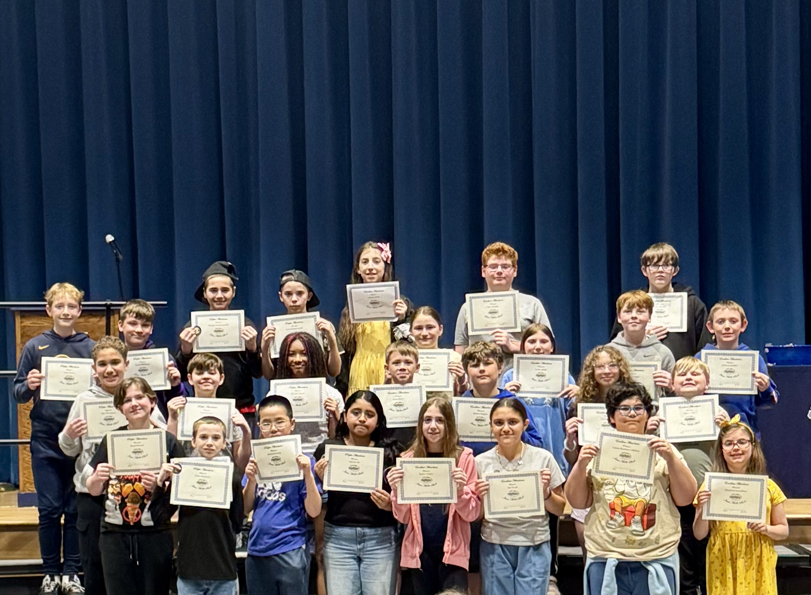 A group of students stand on risers, holding certificates in front of a blue curtain.