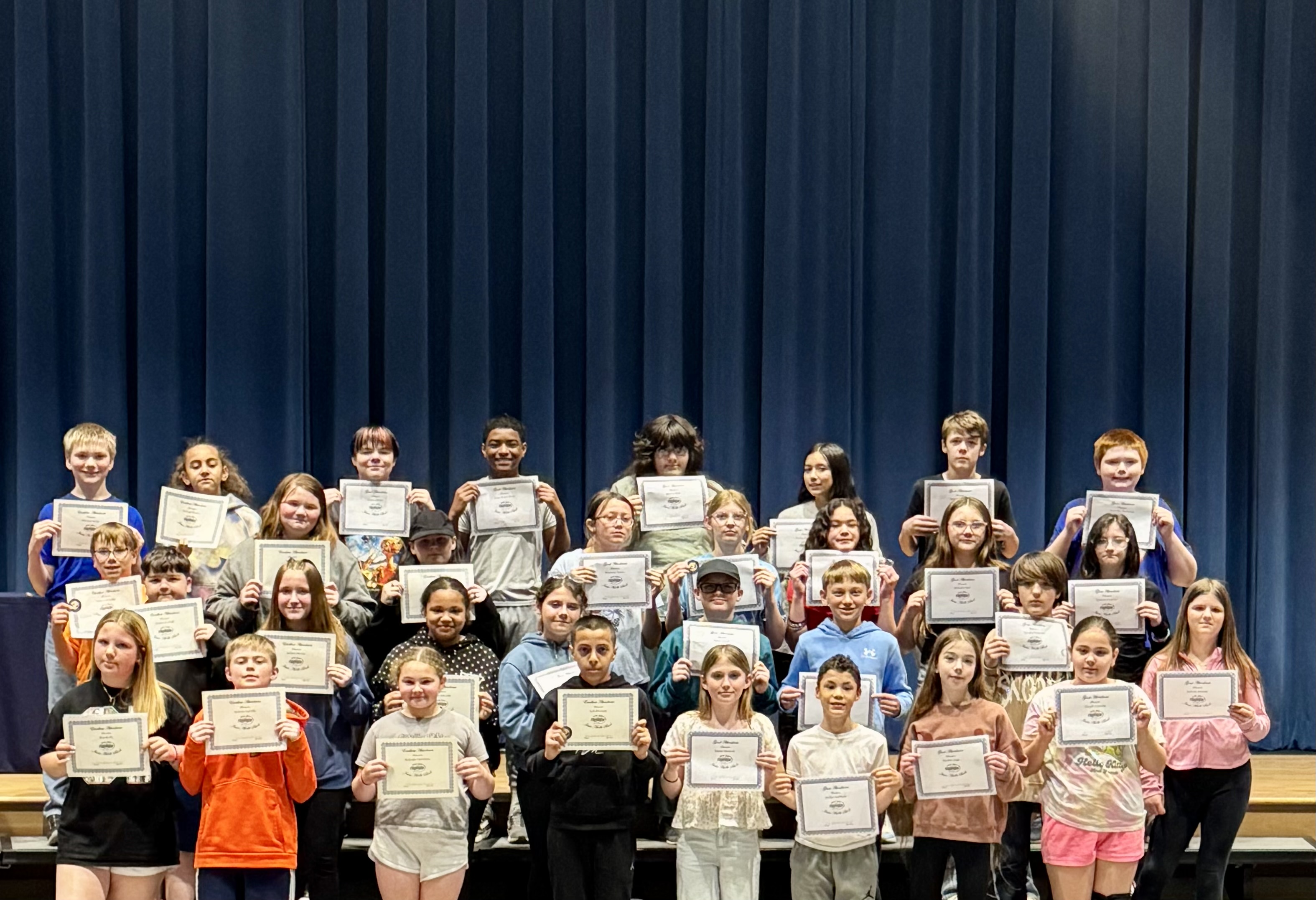 A group of students stand on a stage, holding certificates in front of a blue curtain.