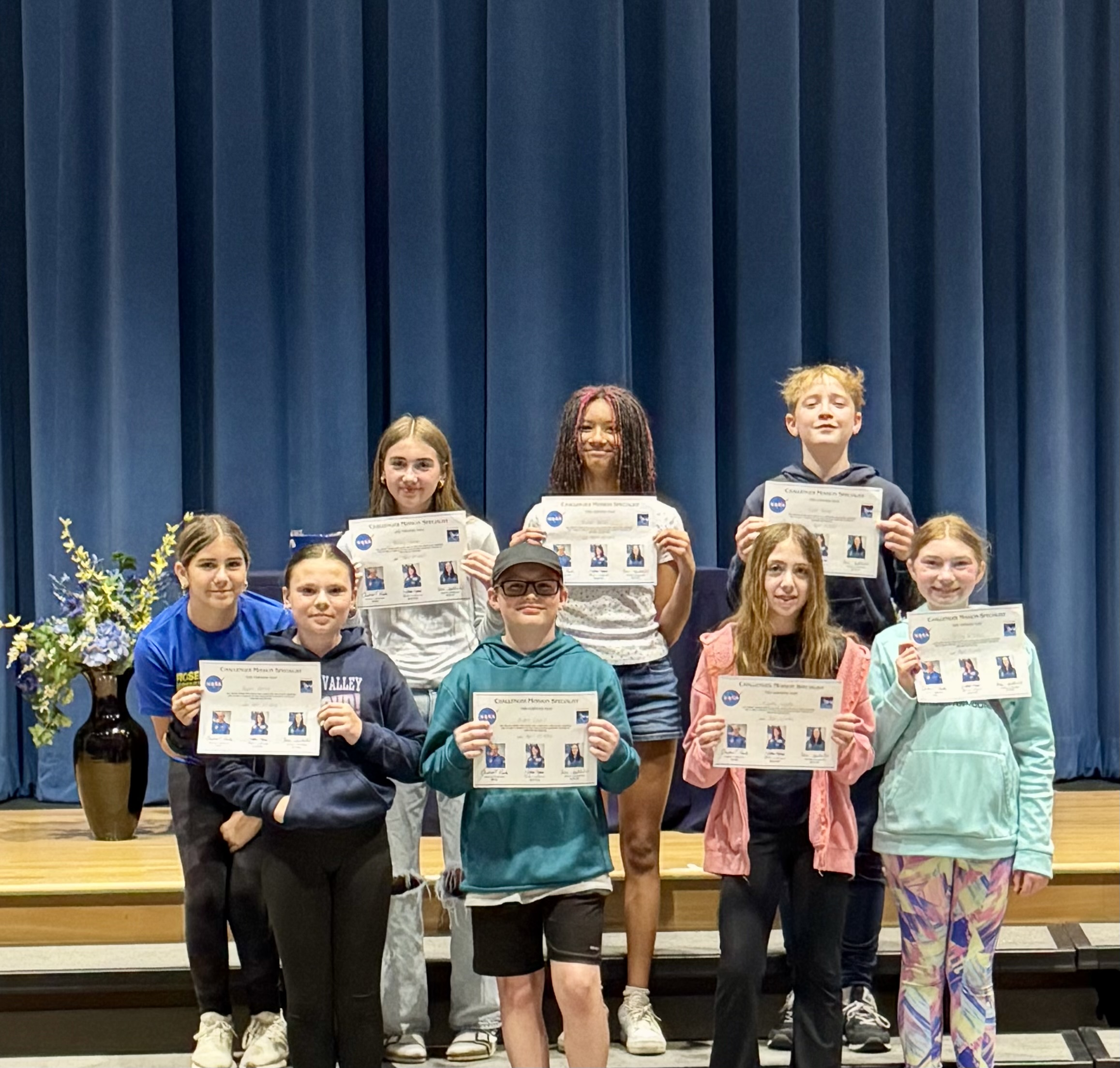Eight students stand on a stage holding certificates, with a blue curtain in the background.
