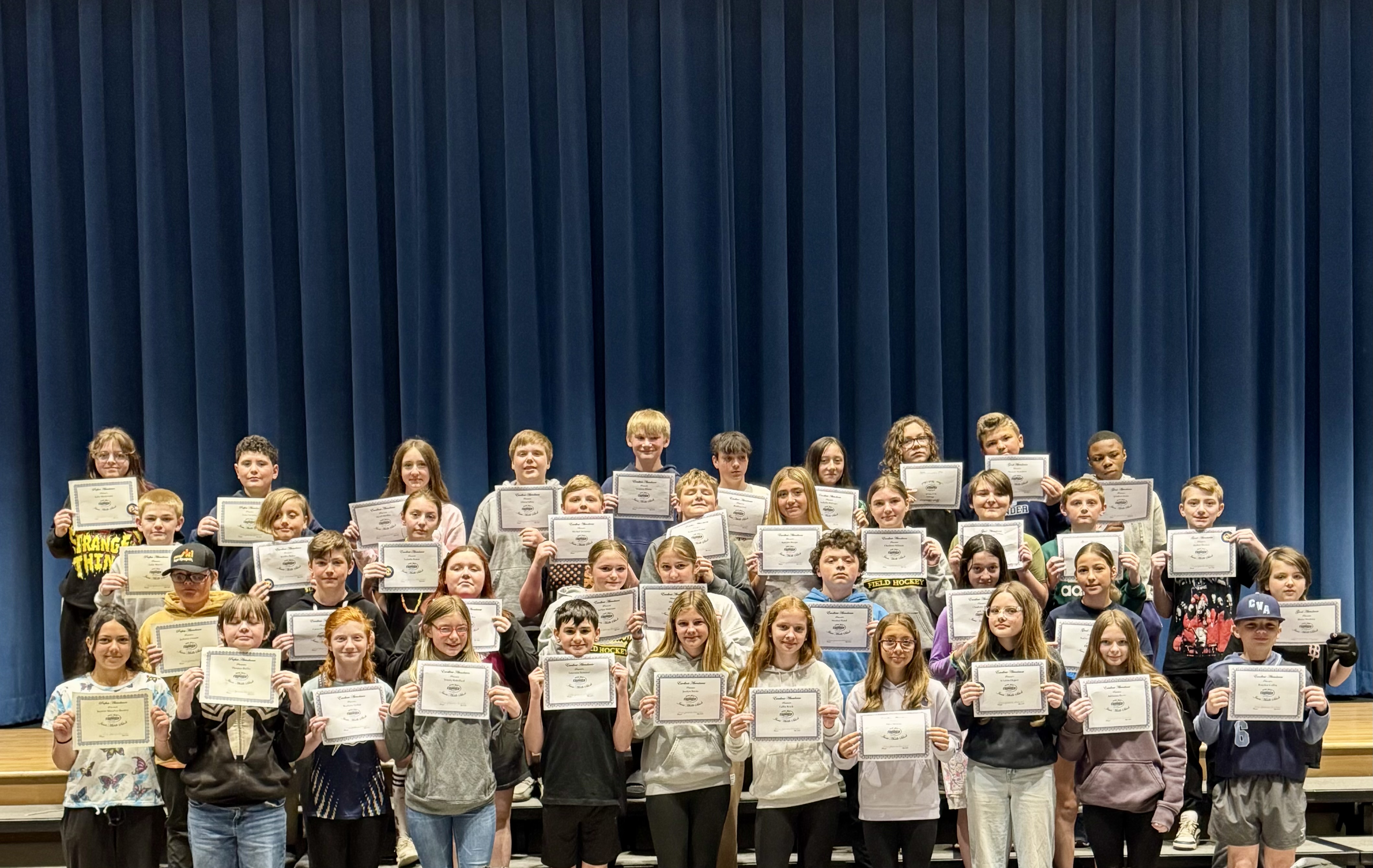 A group of students stand on risers, holding certificates in front of a blue curtain.