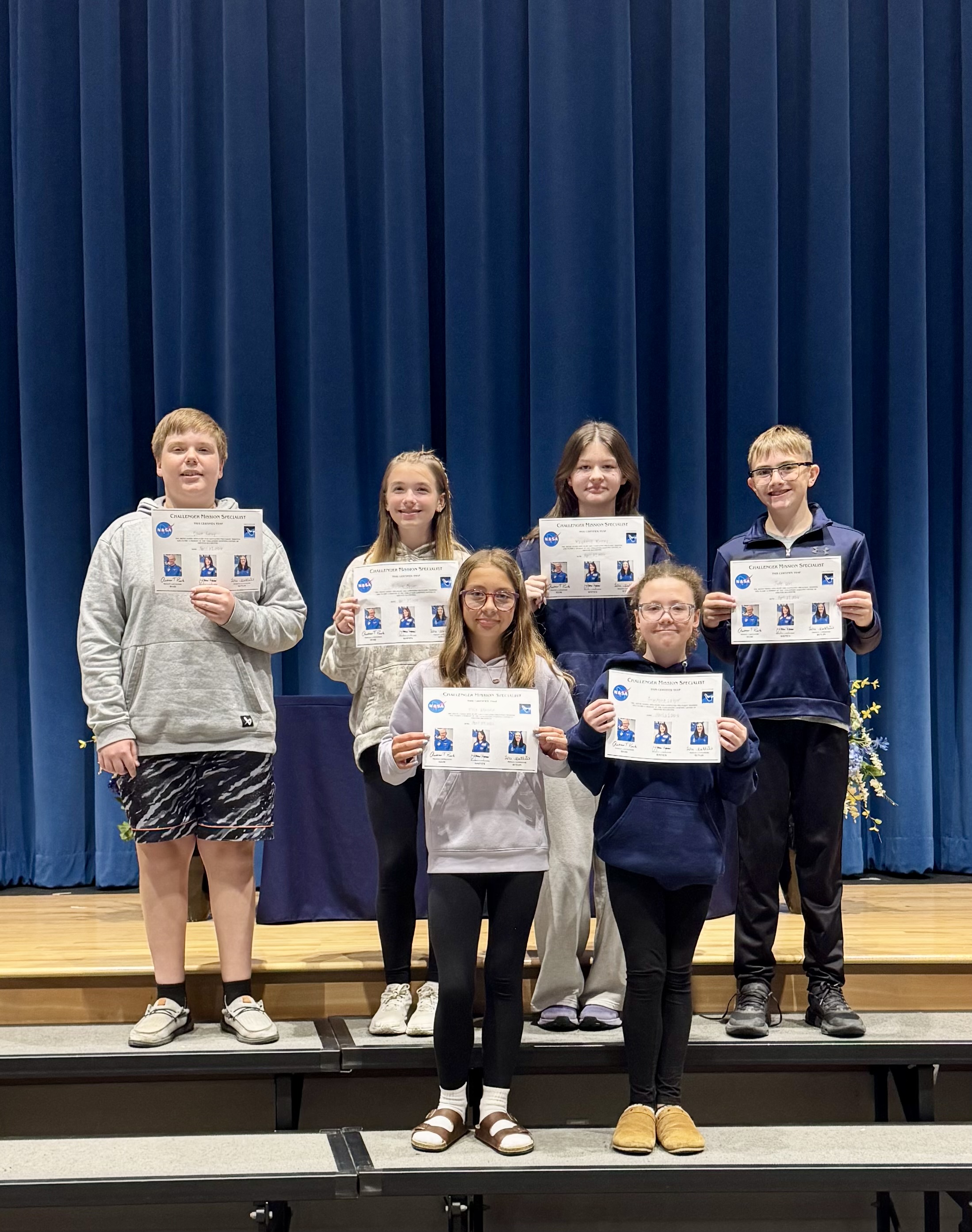 Six students stand on risers, holding certificates that read 'CHALLENGER MISSION SPECIALIST' with NASA logos.